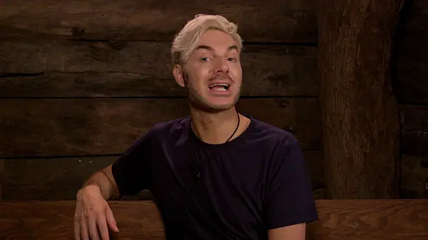 Sam Dylan sitzt in einer Holzhütte und spricht. Er trägt ein dunkles T-Shirt und hat blond gefärbte Haare - Foto:  RTL (DE)