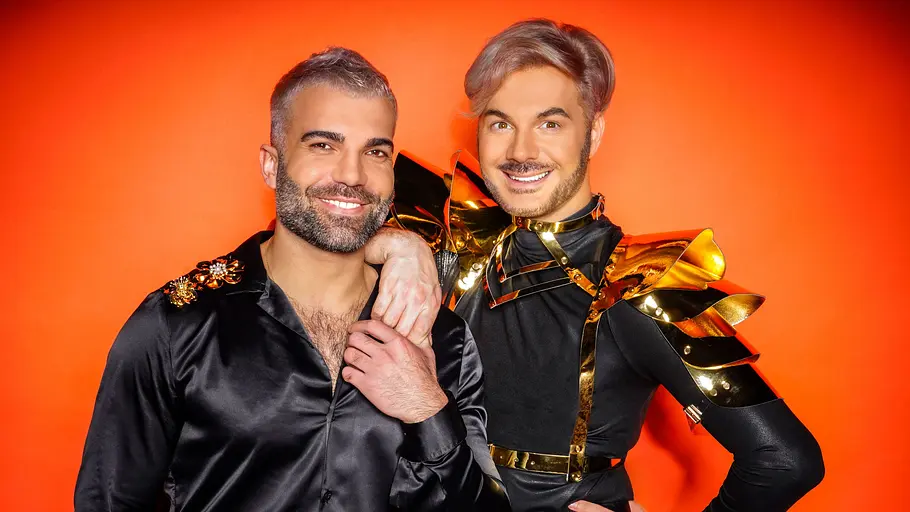 Sam Dylan und Rafi Rachek Sam Dylan und Rafi Rachek posieren lächelnd in schwarzen Outfits mit goldenen Verzierungen vor orangefarbenem Hintergrund.