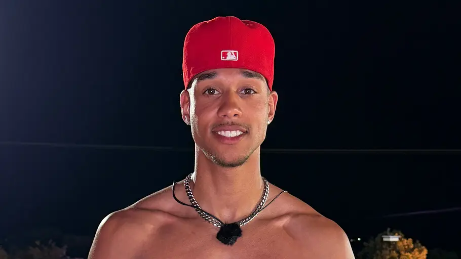 Samuel, 26 Ein Mann mit freiem Oberkörper, roter Kappe und grauer Hose steht nachts vor einem beleuchteten Pool und lächelt.