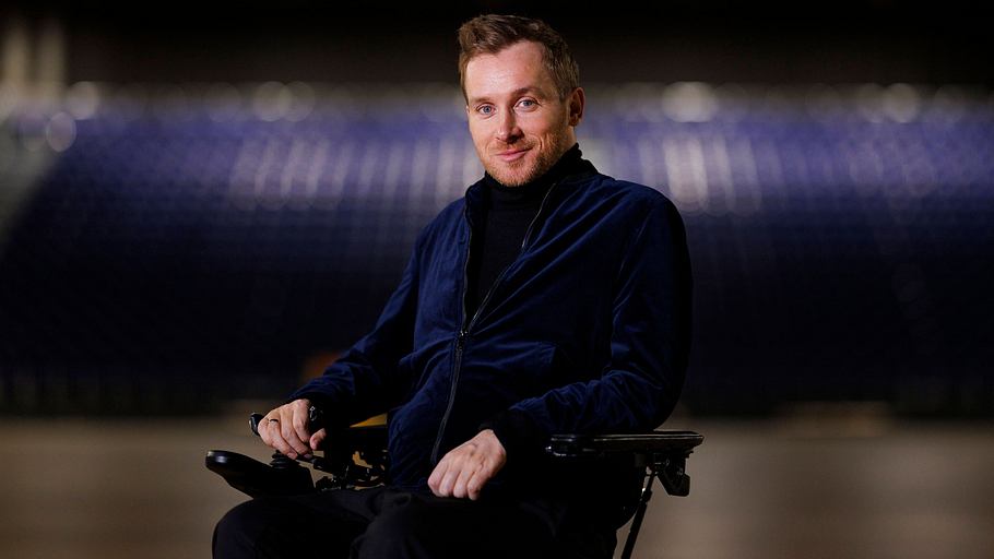 Samuel Koch im Rollstuhl und in blauem Samtblazer in einem Theatersaal