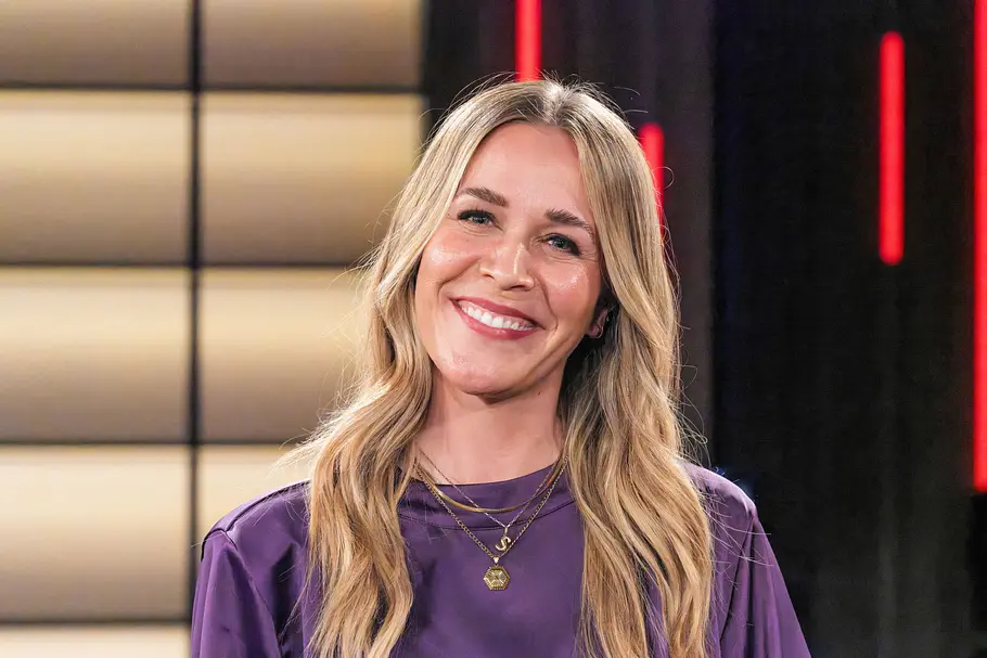 Sandra Sprünken Sandra Sprünken lächelt in die Kamera. Die Moderatorin trägt ein lilafarbenes Oberteil und mehrere goldene Halsketten, ihre langen blonden Haare fallen in sanften Wellen über die Schultern. Im Hintergrund ist ein TV-Studio mit roten Lichtelementen zu sehen.