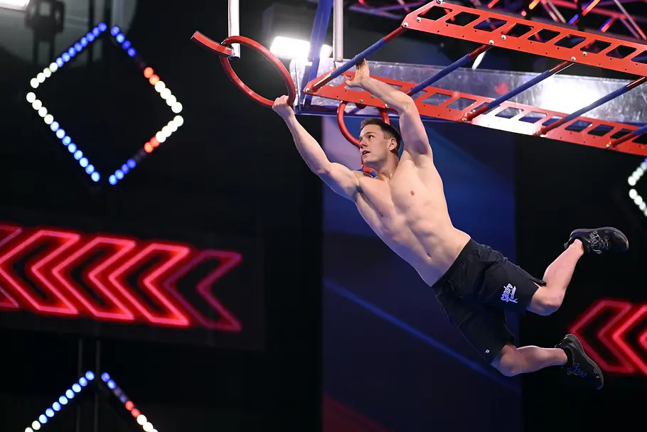 Sandro Scheibler ist Teilnehmer bei Ninja Warrior Germany 2026 Sandro Scheibler schwingt mit freiem Oberkörper an einem roten Ring durch einen beleuchteten Hindernisparcours.