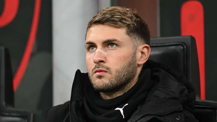 Ein Fußballspieler sitzt mit ernstem Blick auf der Ersatzbank. Er trägt eine schwarze Winterjacke und einen schwarzen Schal, auf seiner Nase klebt ein hautfarbenes Pflaster. Im Hintergrund sind die Sitze der Trainerbank und unscharfe Stadionelemente zu sehen.