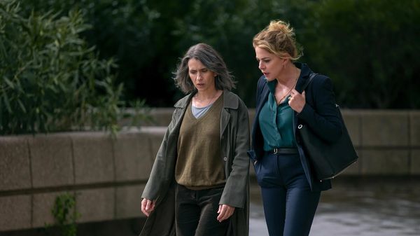 Man sieht Teresa Saponangelo und Claudia Gerini, die auf einer Straße vor einer zugemauerten Grünanlage stehen. Saponangelo trägt eine schwarze Hose ein beiges Oberteil und eine graue Jacke, während Gerini eine dunkelblaue Jeans, eine dunkelgrüne Bluse, sowie eine dunkelblaue Jacke und eine schwarze Handtasche trägt. - Foto: Netflix