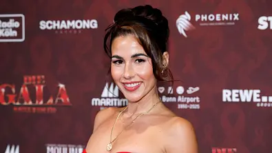 Brünette Frau in einem schulterfreien roten Kleid lächelt auf einem roten Teppich vor einer Event-Fotowand mit Sponsor-Logos in die Kamera. - Foto: IMAGO / Panama Pictures