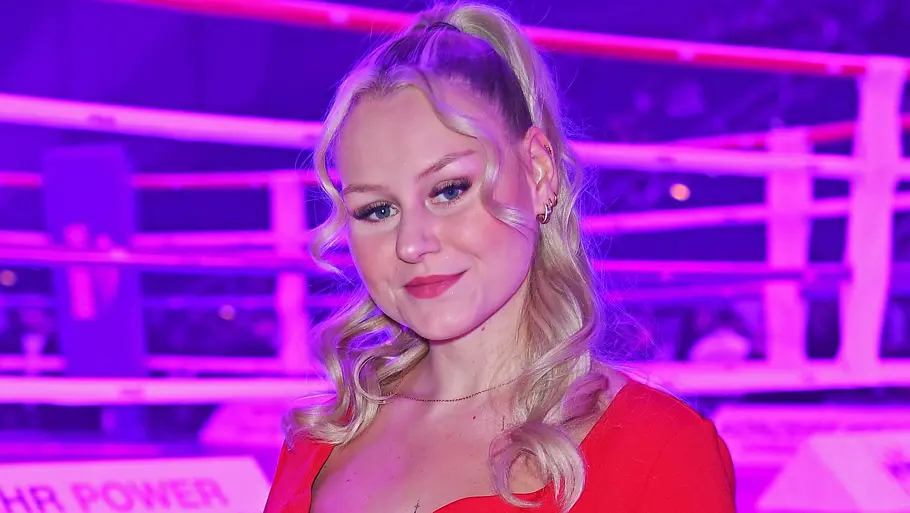 Sarah-Jane Wollny steht bei einer Veranstaltung vor einem beleuchteten Boxring und trägt ein rotes Kleid