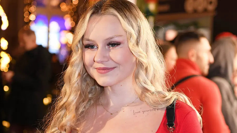 Sarah-Jane Wollny, die aktuell ihre Lipödem-Diagnose verdaut, lächelt bei einem Event in rotem Outfit vor unscharfem Hintergrund.