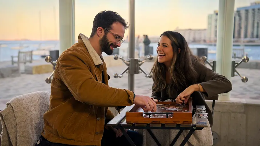 Sarah und John. Sarah und John sitzen lachend an einem kleinen Tisch mit einem Puzzle. Hinter großen Fenstern liegt eine Uferpromenade.