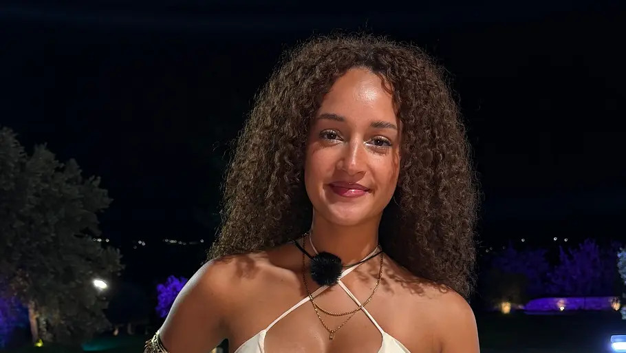 Saskia H., 27 Eine Frau mit langen Locken und goldfarbenem Schmuck steht nachts in einem cremefarbenen Zweiteiler an einem beleuchteten Pool.