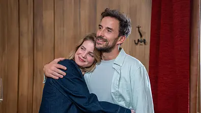 Eine Frau in Jeansjacke und ein Mann in hellblauem Hemd umarmen sich lächelnd vor einer Holzwand. Die beiden gehören zum Ensemble der Serie Die Landarztpraxis. - Foto: Joyn / Joshua Schneider Eine Frau in Jeansjacke und ein Mann in hellblauem Hemd umarmen sich lächelnd vor einer Holzwand. Die beiden gehören zum Ensemble der Serie Die Landarztpraxis. - Foto: Joyn / Joshua Schneider