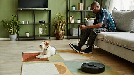 Ein Saugroboter dreht seine Runden während rechts ein Mann auf der Couch chillt und Mitte links liegt ein Hund auf dem Teppich. - Foto: iStock / SeventyFour 