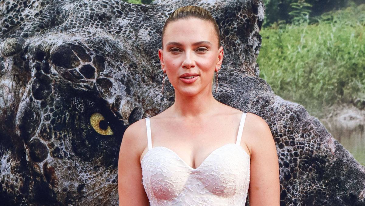 Hatte Scarlett Johansson überhaupt „Jurassic World 4“-Konkurrenz? Scarlett Johansson steht in einem weißen Kleid vor einem „Jurassic World: Die Wiedergeburt“-Plakat und lächelt verschmitzt