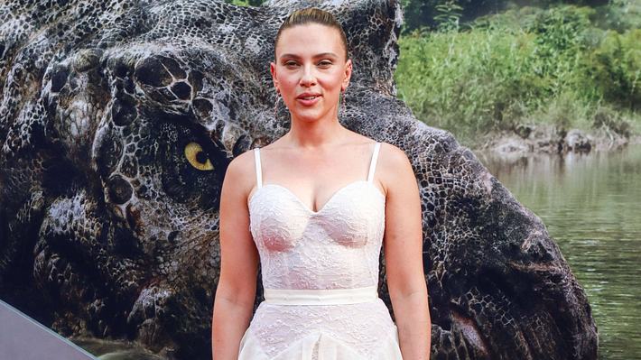 Scarlett Johansson steht in einem weißen Kleid vor einem „Jurassic World: Die Wiedergeburt“-Plakat und lächelt verschmitzt - Foto:  IMAGO / ABACAPRESS