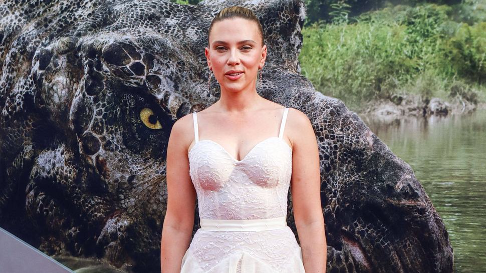 Scarlett Johansson steht in einem weißen Kleid vor einem „Jurassic World: Die Wiedergeburt“-Plakat und lächelt verschmitzt - Foto:  IMAGO / ABACAPRESS