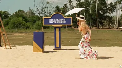Dolly Buster läuft über einen sandigen Platz an einem Schild mit der Aufschrift „Die goldene Mitte finden“ vorbei; im Hintergrund sind Palmen zu sehen. - Foto: Joyn