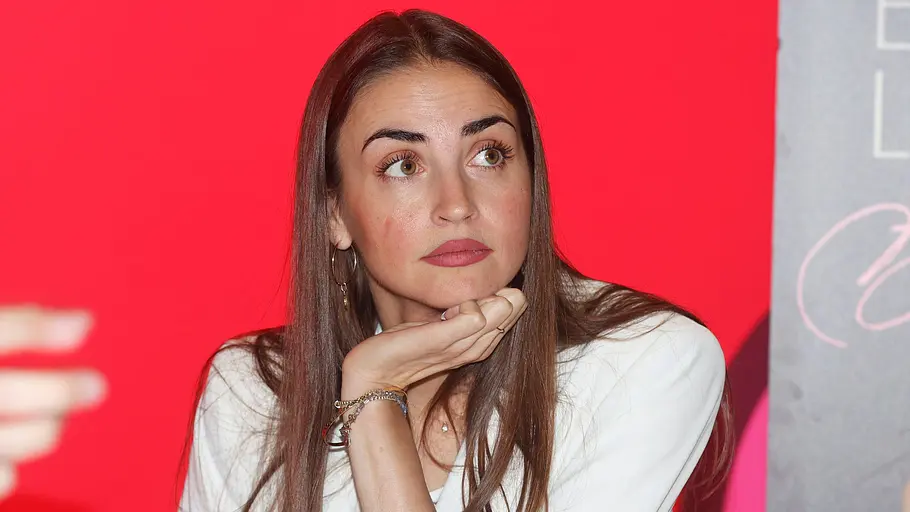 Schwere Nachrichten für Ekaterina Leonova Ekaterina Leonova sitzt in weißer Bluse vor rotem Hintergrund an einem Tisch und stützt das Kinn auf ihre Hand.