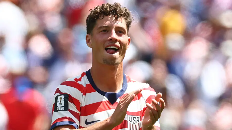 Ein US-amerikanischer Fußballspieler im rot-weiß gestreiften Nationaltrikot applaudiert auf dem Spielfeld. Mit geöffnetem Mund blickt er in Richtung Tribüne, während der Hintergrund mit den Zuschauern unscharf verschwimmt.