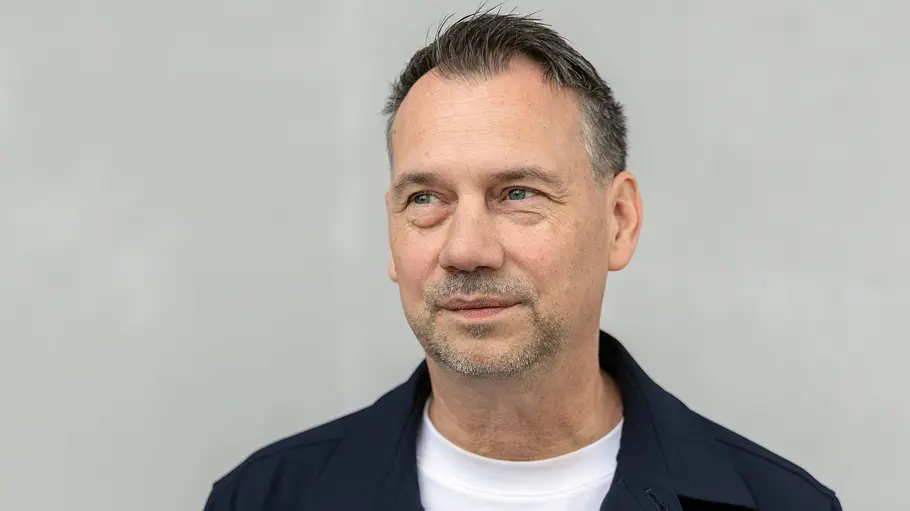 Sebastian Fitzek Sebastian Fitzek trägt eine dunkelblaue Jacke über einem weißen Shirt und blickt vor grauem Hintergrund zur Seite.