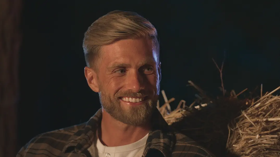 Sebastian Paul aus „Die Bachelors“ lächelt bei einem Abenddate vor Heuballen im Hintergrund. - Foto: RTL