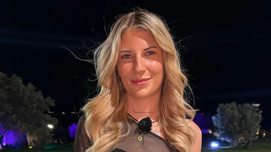 Selina, 20 Eine blonde Frau mit Tattoos trägt ein schwarzes Kleid mit transparentem Oberteil und steht nachts vor einem beleuchteten Pool.