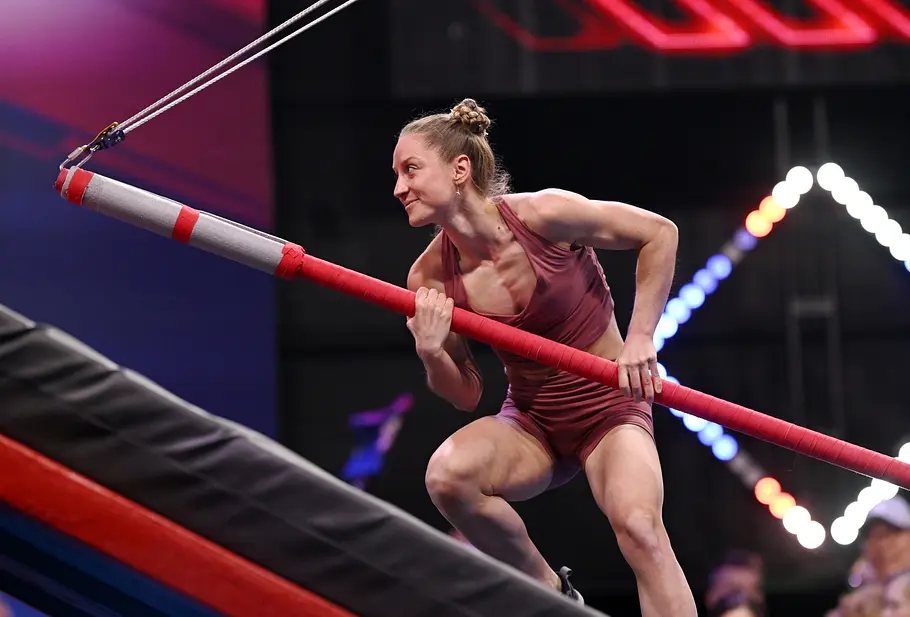 Selina Schwemmer ist Teilnehmer bei Ninja Warrior Germany 2026 Selina Schwemmer hält in bordeauxroter Sportkleidung eine schräg hängende, rot umwickelte Stange über einer gepolsterten Rampe.