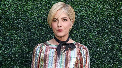 Selma Blair steht mit blonden kurzen Haaren, buntem Paillettenkleid und schwarzer Halsschleife vor einer grünen Hecke. - Foto:  IMAGO / Image Press Agency