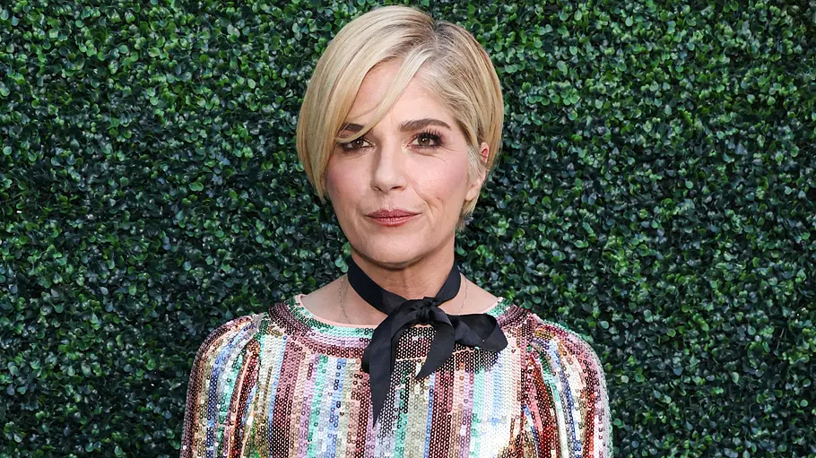 Selma Blair Vorfall Flugzeug Angriff Selma Blair steht mit blonden kurzen Haaren, buntem Paillettenkleid und schwarzer Halsschleife vor einer grünen Hecke.