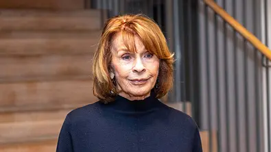 Senta Berger trägt einen Pullover und steht vor einer Treppe. - Foto: IMAGO / Hartenfelser