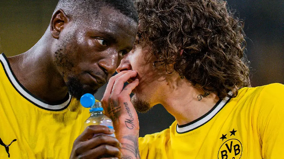 BVB-Stürmer Guirassy hält eine Trinkflasche, während ein BVB-Mitspieler im gelben Heimtrikot etwas zuflüstert.