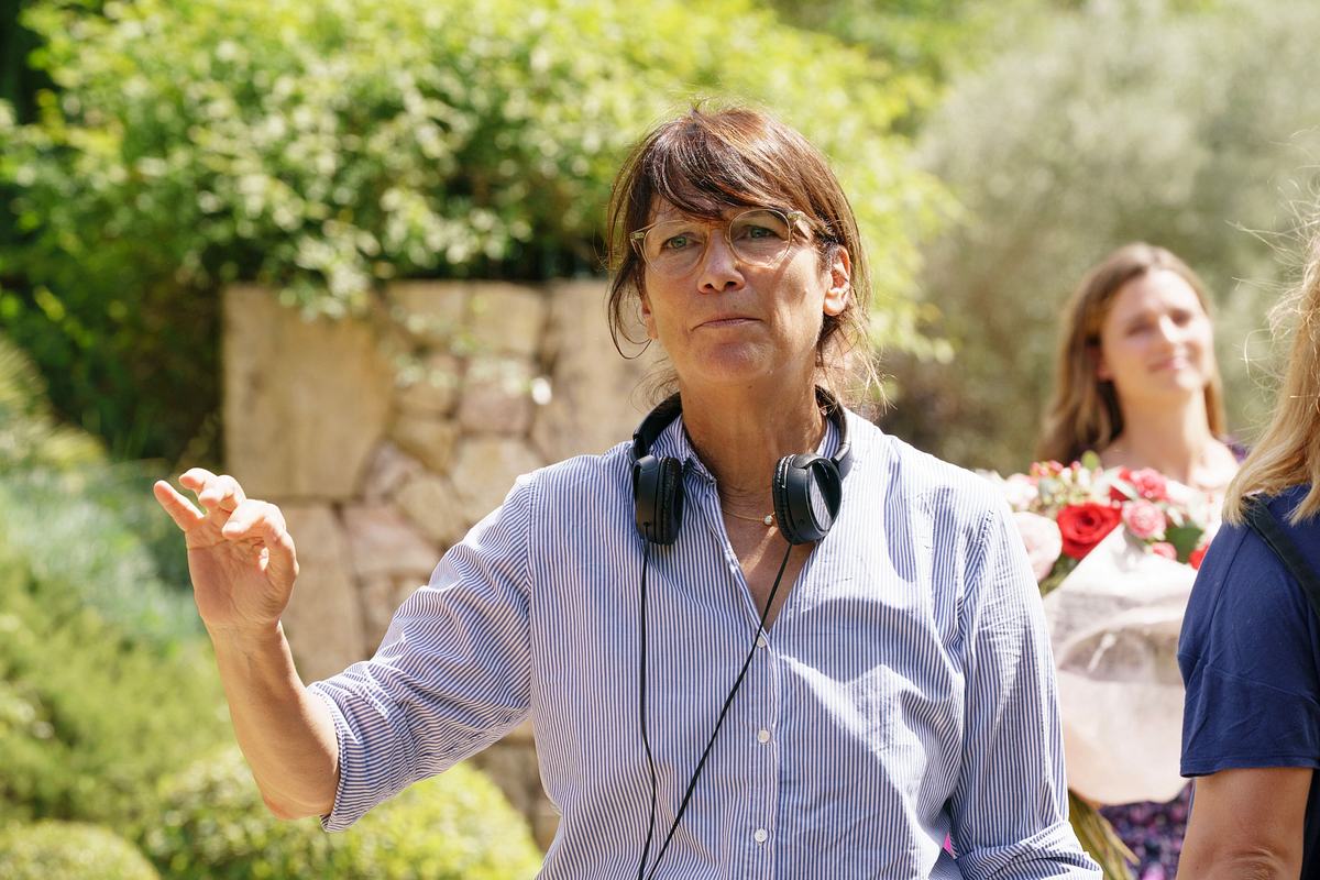 Auf dem Foto ist Regisseurin Sherry Hormann zu sehen. Sie steht im Freien vor einer Steinmauer und grünen Pflanzen. Sie trägt eine hellblau-weiß gestreifte Bluse, eine Brille und hat ein schwarzes Headset um den Hals hängen. Mit der rechten Hand macht sie eine erklärende Geste, als würde sie gerade etwas am Filmset anweisen.