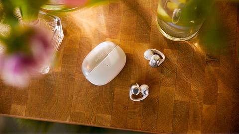 Die Shokz OpenDots ONE Clip-On-Ohrhörer liegen auf einem Holztisch, links und rechts davon Blumenvasen, die unscharf nicht im Fokus der Aufnahme sind. - Foto: ©2025 Shokz