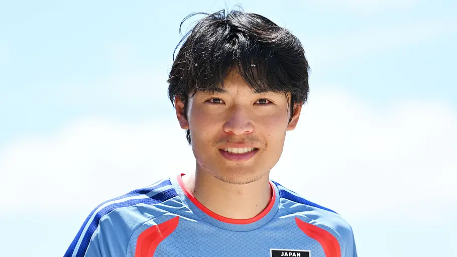 Fußballspieler der japanischen Nationalmannschaft in einem hellblauen Trainingstrikot blickt lächelnd in die Kamera. Im Hintergrund sind ein heller Himmel und unscharfe Elemente im Freien zu sehen.