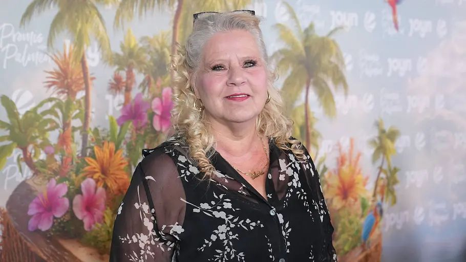Silvia Wollny steht vor eine Fotowand der Sendung „Promis unter Palmen“. 