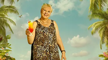 Eine ältere Frau in einem luftigen, ärmellosen Kleid mit Leopardenmuster steht barfuß im Sand vor einer tropischen Strandkulisse mit Palmen, bunten Blumen und türkisfarbenem Meer. In der Hand hält sie einen fruchtigen Cocktail mit Kirsche und Orangenscheibe, während über ihr bunte Papageien fliegen. - Foto: Joyn