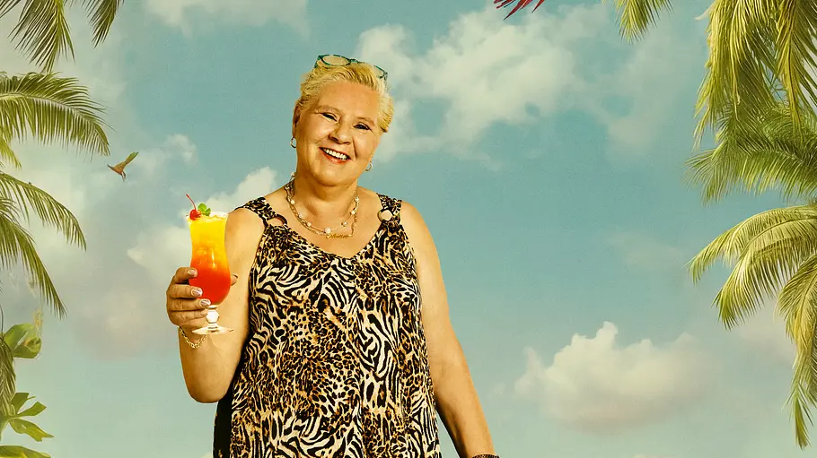 Eine ältere Frau in einem luftigen, ärmellosen Kleid mit Leopardenmuster steht barfuß im Sand vor einer tropischen Strandkulisse mit Palmen, bunten Blumen und türkisfarbenem Meer. In der Hand hält sie einen fruchtigen Cocktail mit Kirsche und Orangenscheibe, während über ihr bunte Papageien fliegen.