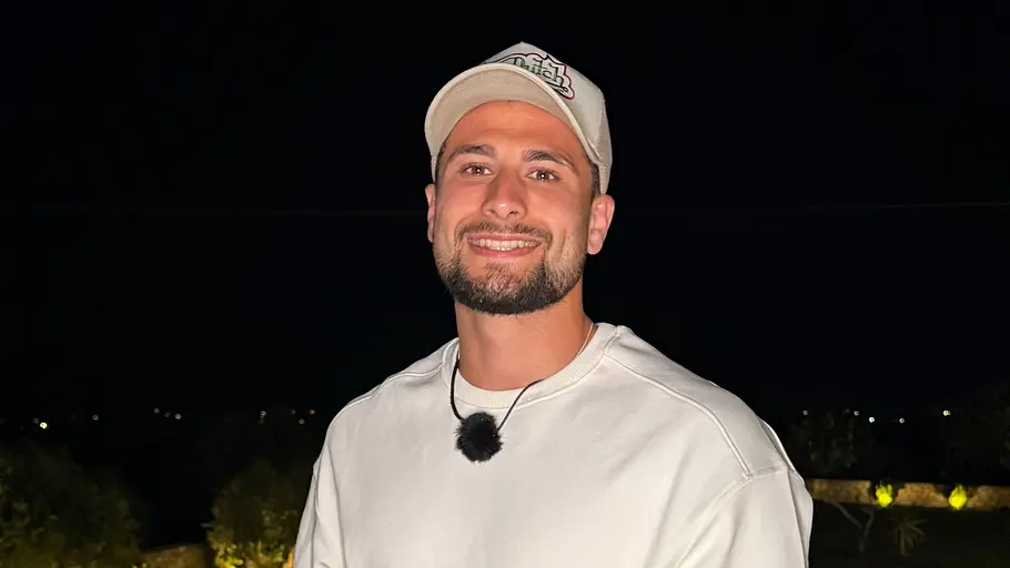 Simon, 25 Ein lächelnder Mann mit Kappe, weißem T-Shirt, rosa Shorts und tätowiertem Arm steht nachts vor einem beleuchteten Pool.