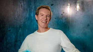Simon Gosejohann steht lächelnd vor einem blau-grau beleuchteten Hintergrund. Er trägt ein helles, langärmeliges Shirt. - Foto: © SWR/Christian Koch