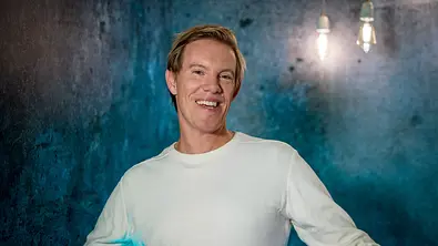 Simon Gosejohann steht lächelnd vor einem blau-grau beleuchteten Hintergrund. Er trägt ein helles, langärmeliges Shirt. - Foto: © SWR/Christian Koch
