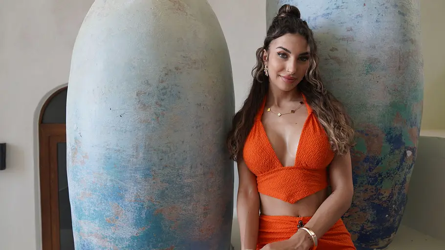 Siria steht in einem orangenen Zweiteiler zwischen zwei Statuen und schaut in die Kamera.