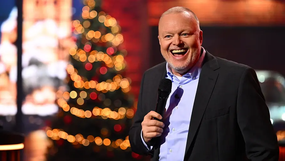 Stefan Raab moderiert lachend vor einem Weihnachtsbaum.