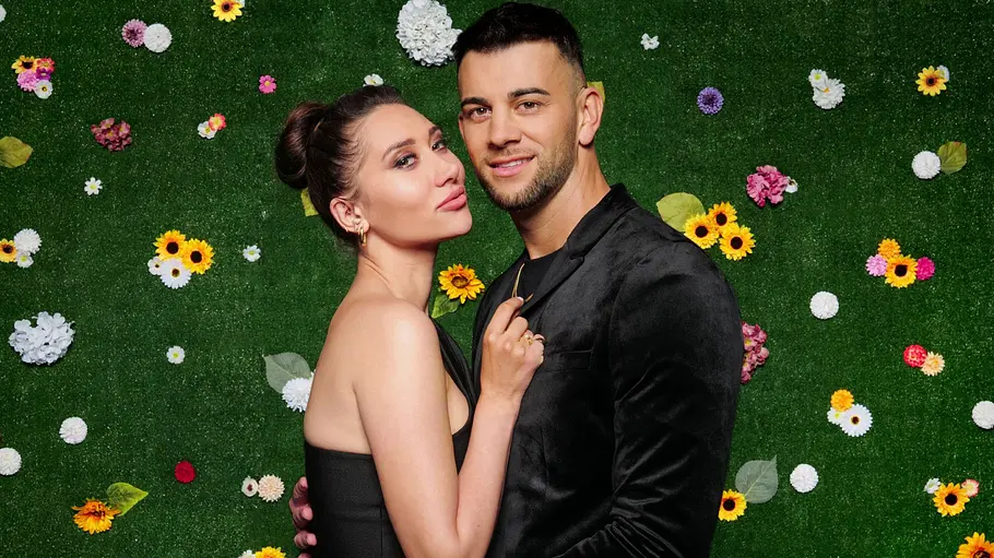 Samira und Serkan Samira und Serkan Yavuz posieren eng umschlungen vor einer grünen Blumenwand. Das Reality-TV-Paar blickt lächelnd in die Kamera und trägt elegante schwarze Outfits.