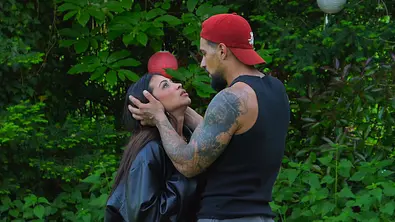 Ein tätowierter Mann mit roter Kappe hält den Kopf einer Frau in seinen Händen. Sie stehen vor dichtem Grün und schauen sich an. - Foto: RTL Ein tätowierter Mann mit roter Kappe hält den Kopf einer Frau in seinen Händen. Sie stehen vor dichtem Grün und schauen sich an. - Foto: RTL