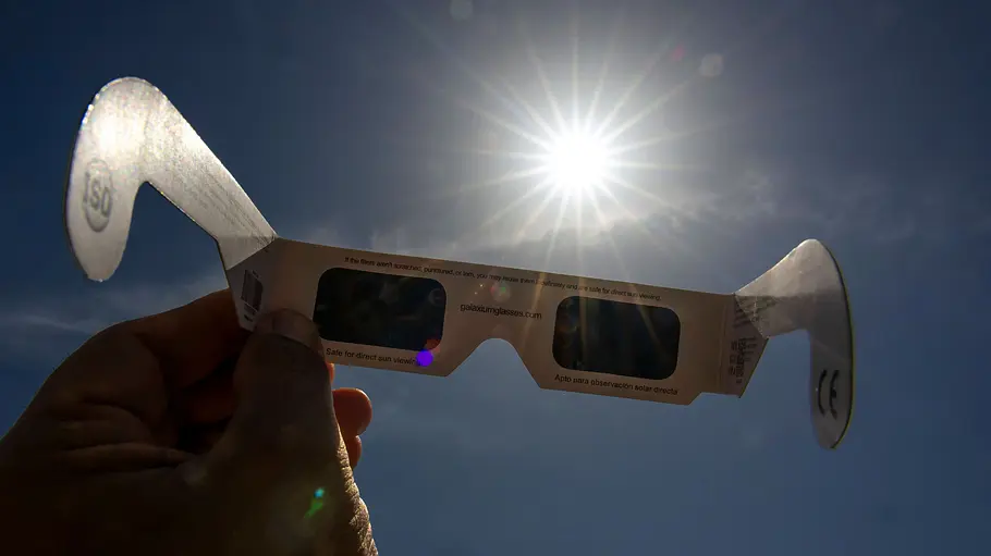 Sonnenfinsternis mit Schutzbrille beobachten Eine Hand hält eine spezielle Sonnenfinsternisbrille vor die hell leuchtende Sonne am Himmel.
