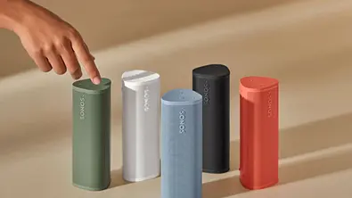 Mehrere Sonos Roam 2 Lautsprecher in verschiedenen Farben stehen nebeneinander auf einer hellen Fläche, während eine Hand die Oberseite eines grünen Modells berührt. Die kompakten WLAN- und Bluetooth-Speaker sind zylindrisch geformt und tragen das Sonos-Logo auf der Front. - Foto: Sonos/PR
