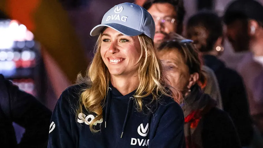 Sophia Flörsch lächelt mit verschränkten Armen, blauer Kappe und dunklem Kapuzenpullover. Hinter ihr stehen mehrere Menschen.