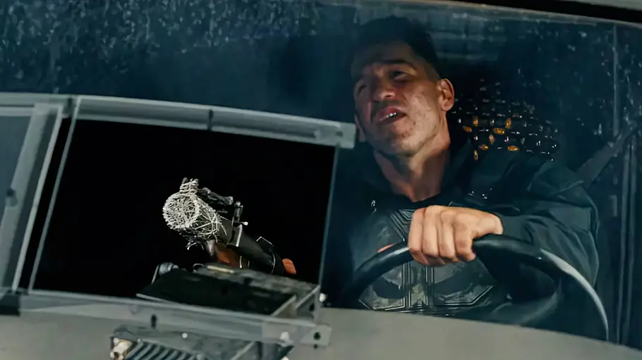 Punisher Jon Bernthal als Punisher sitzt am Steuer eines Fahrzeugs, hält eine Waffe in der Hand und blickt während einer nächtlichen Verfolgungsjagd entschlossen nach vorn.