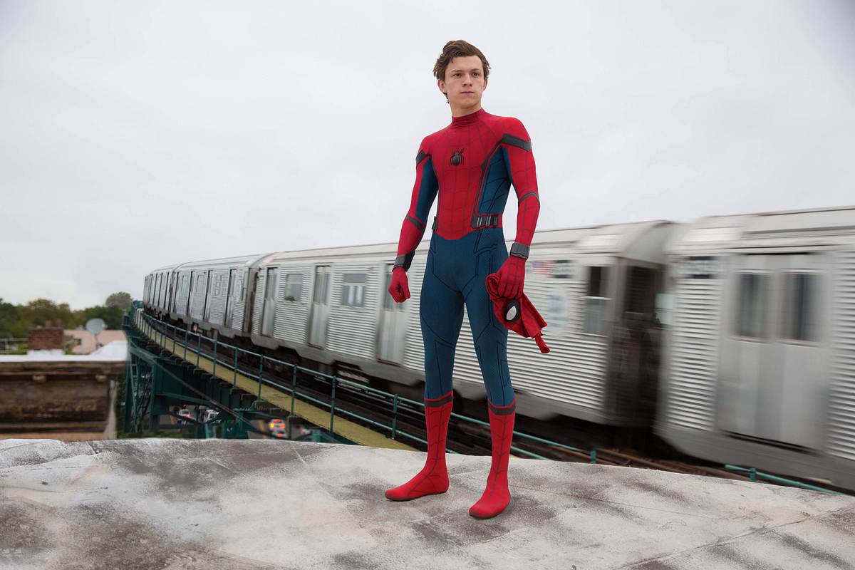 Tom Holland Tom Holland steht als Spider-Man auf einem Plateau und hinter ihm fährt ein Zug vorbei. Er trägt keine Maske.