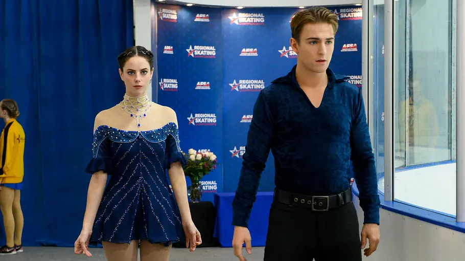 Netflix-Serie „Spinning Out“ Ein Eiskunstlaufpaar steht nebeneinander am Rand einer Eishalle, bereit zum Betreten der Eisfläche. Die Frau trägt ein dunkelblaues, glitzerndes Eiskunstlaufkleid und Schlittschuhe, der Mann ein dunkelblaues Oberteil und schwarze Hose. Im Hintergrund sind weitere Sportlerinnen und Sportler sowie ein blauer Vorhang und eine Wand mit Logos zu sehen.