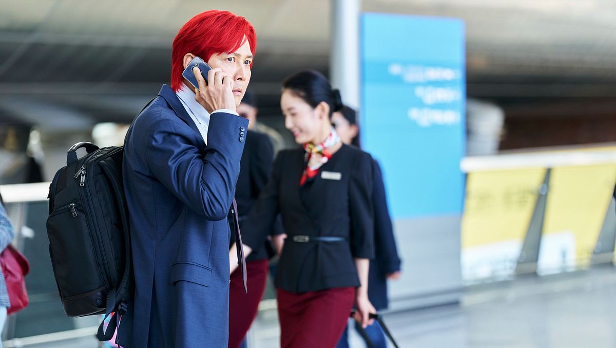 Squid Game Gi-hun steht mit roten Haaren am Flughafen. Er telefoniert und hat dabei einen ernsten Gesichtsausdruck