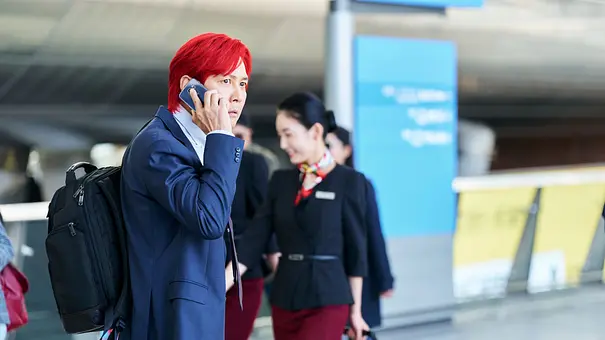 Gi-hun steht mit roten Haaren am Flughafen. Er telefoniert und hat dabei einen ernsten Gesichtsausdruck - Foto: No Ju-han/Netflix Gi-hun steht mit roten Haaren am Flughafen. Er telefoniert und hat dabei einen ernsten Gesichtsausdruck - Foto: No Ju-han/Netflix